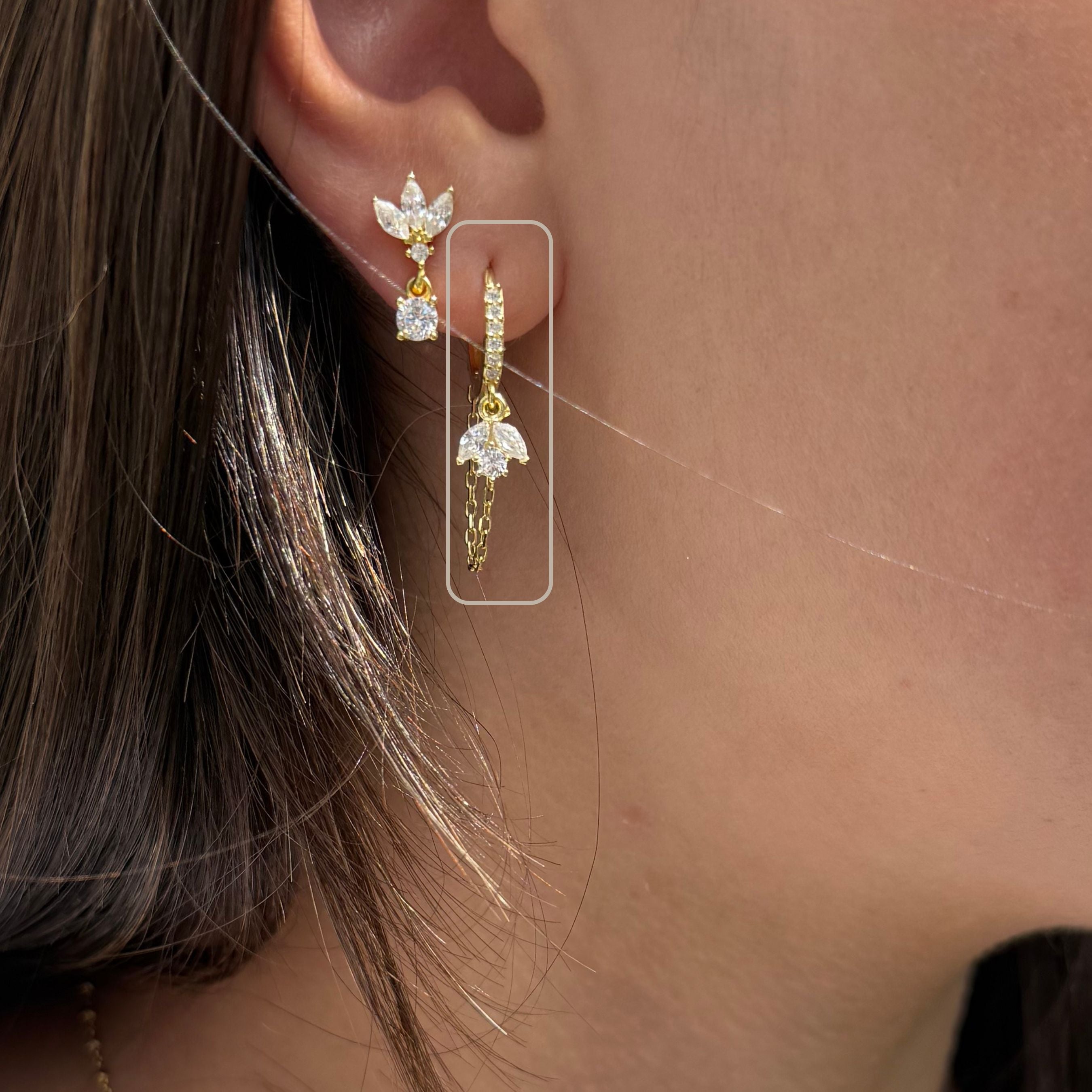 Close-up van gouden oorbel met bloemvormige zirkonia hanger en fijn kettinkje, gedragen in het oor
